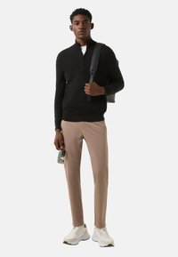 Zwarte trui met rits, lichtbeige broek en witte sneakers. Houdt een zonnebril vast, draagt een zwarte schoudertas, staand tegen een effen achtergrond.