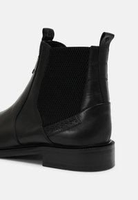 Černá kožená Chelsea bota s texturovanou elastickou boční částí, poutky na patě a kotníku a zúženým podpatkem na jednobarevném pozadí.