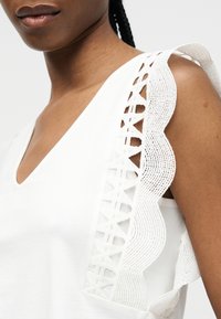 Γυναίκα που φοράει λευκό αμάνικο τοπ με V-λαιμόκοψη και υφασμάτινη δαντελένια βολάν λεπτομέρεια στον ώμο, σε μονόχρωμο φόντο.