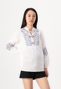 Blusa bianca con motivi floreali blu ricamati, colletto arricciato e maniche lunghe a sbuffo. Abbinata a pantaloni corti neri.
