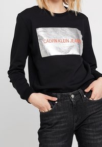 Felpa nera con un pannello rettangolare argentato metallico con "CALVIN KLEIN JEANS" in bold rosso. Tessuto morbido, maniche lunghe.