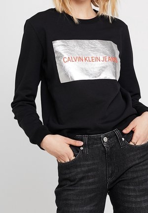 Svart sweatshirt med en metallisk silver rektangulär panel med "CALVIN KLEIN JEANS" i fet röd text. Mjuk tyg, långa ärmar.