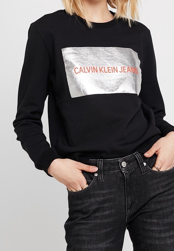Felpa nera con un pannello rettangolare argentato metallico con "CALVIN KLEIN JEANS" in bold rosso. Tessuto morbido, maniche lunghe.