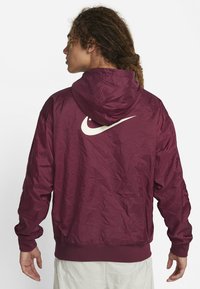 Bordo rdeča jakna Nike z gubano teksturo, kapuco in belim logotipom na hrbtni strani. Ima elastične manšete in pas za trden oprijem.