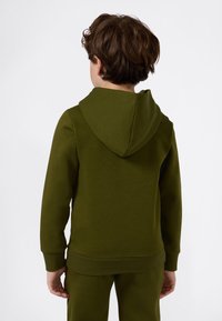 Bambino con capelli ricci castani che indossa un hoodie verde oliva e pantaloni abbinati, in piedi rivolto di spalle contro uno sfondo bianco uniforme.