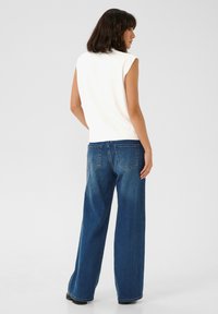 Top blanc sans manches associé à un jean bleu foncé ample avec un design à cinq poches et une texture délavée, vu de dos.