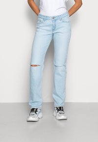 Parecias de ganga azul claro com perna reta, apresentando um único rasgo no joelho e um clássico design de cinco bolsos. Usadas com sapatilhas brancas e cinzas.