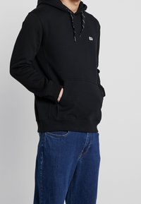 Sweat à capuche noir avec poche avant, arborant un petit logo blanc. Accompagné d'un jean bleu, vu de profil.