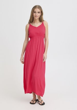 Jeune femme portant une robe maxi rose sans manches et des tongs noires, debout devant un fond blanc uni.