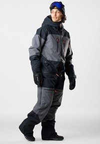 Man wearing a black and gray ski suit, black gloves, and blue ski goggles, standing with one leg slightly forward looking to the side.