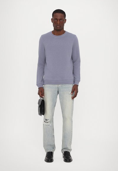 Un pull crewneck violet clair, un jean bleu clair usé, des chaussures noires et un petit sac noir porté à une main.