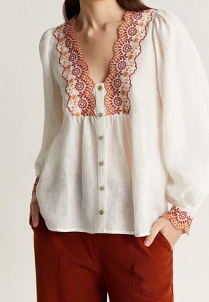 Biała guziczka z balonowymi rękawami, zdobiona pomarańczowo-czerwonym haftem kwiatowym z falistym brzegiem na kołnierzu i mankietach, noszona z rdzawymi spodniami.