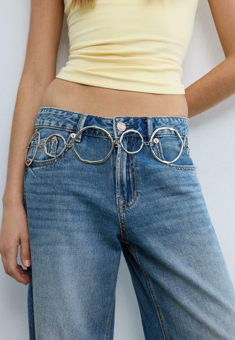 Frau trägt blaue Jeans mit einem silbernen, geschlungenen Ketten-Gürtel und einem blassgelben, bauchfreien Oberteil, steht vor einer weißen Wand.