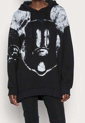 Czarna oversize bluza z kapturem z abstrakcyjną białą grafiką przypominającą zniszczoną twarz kreskówkową, noszona przez osobę stojącą na jednolitym tle.