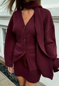 Burgundský pletený kardigan s výstřihem do V, zapínáním na knoflíky a širokými rukávy, zkombinovaný s odpovídající pletenou mini sukní a šálou. Hladká textura.