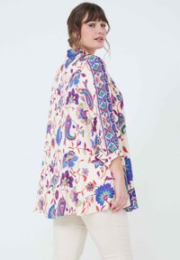 Flowy blouse in cream with vibrant floral and paisley patterns in blue, purple, pink, and green; features buttoned sleeves and detailed trim.