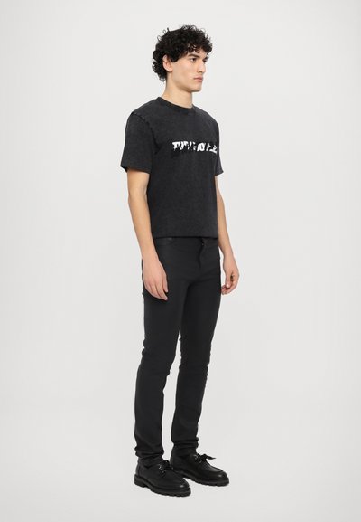 Zwart katoenen t-shirt met een witte grafische afdruk, gecombineerd met zwarte slim-fit broeken en zwarte veterschoenen, staande tegen een witte achtergrond.