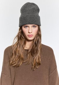 Beanie grigio in lana con una trama a coste, con una leggera caduta. Abbinato a un maglione marrone in maglia texturizzata.