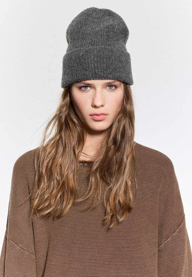 Beanie grigio in lana con una trama a coste, con una leggera caduta. Abbinato a un maglione marrone in maglia texturizzata.