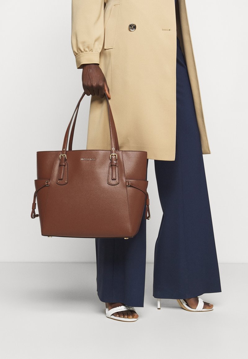 Personne tenant un grand sac à main en cuir marron, portant un manteau beige, un pantalon large bleu marine et des sandales blanches à talons sur fond clair.