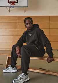 Hanorac negru și pantaloni de trening cu accente aurii. Material texturat, croială relaxată. Adidași argintii cu detalii din plasă, așezați pe o bancă din lemn.