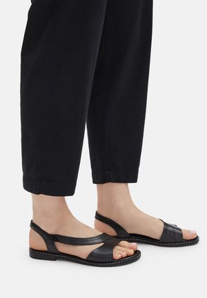 Černé kožené sandály s vyřezávaným designem, otevřenou špičkou a páskem kolem kotníku. Mají šitou podrážku s malými cvočky podél okraje.