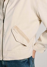 Veste bomber beige avec fermeture éclair à l'avant, présentant une poche diagonale avec le logo "Pepe Jeans", un tissu texturé et des poignets élastiques.