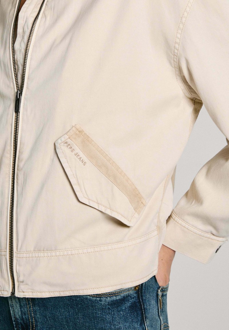 Veste bomber beige avec fermeture éclair à l'avant, présentant une poche diagonale avec le logo "Pepe Jeans", un tissu texturé et des poignets élastiques.
