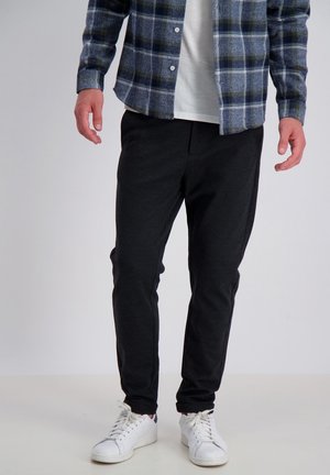 Schwarze Jogginghosen mit schmaler Passform, aus weichem Material, kombiniert mit einem blauen karierten Hemd mit Knopfleiste und weißen Sneakers.