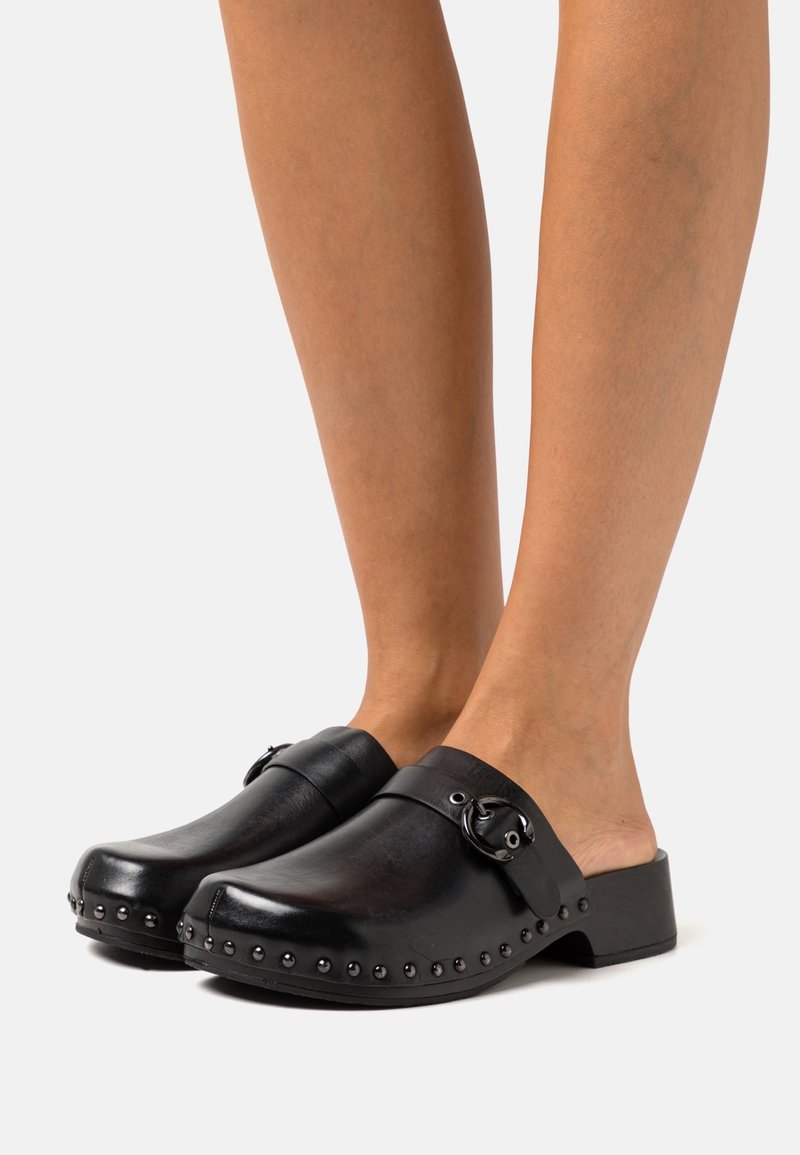 Jambes portant des sabots en cuir noir sans lacets, avec des clous ronds le long du bord de la semelle et une boucle décorative sur le côté.
