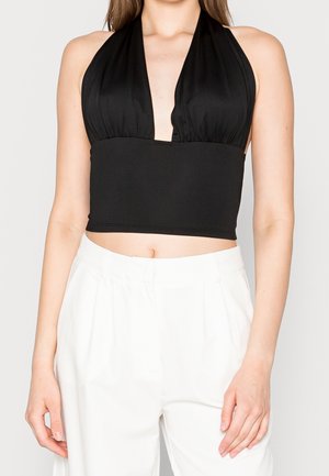 Kvinde iført en sort ærmeløs halter crop top med dyb V-hals, kombineret med højtaljede hvide bukser mod en ensfarvet baggrund.