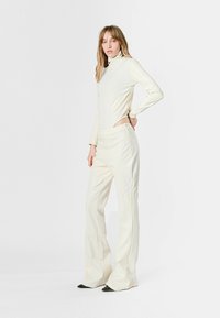 Top ajustado color crema con cuello alto y cortes laterales, combinado con pantalones acampanados. La tela tiene un acabado suave con detalles de costura y tacones negros.