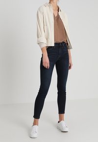 Helles beiges Cordjackett über einem braunen V-Ausschnitt-Oberteil, kombiniert mit dunkelblauen Skinny Jeans und wei�ßen Sneakers. Schlichtes Design und Farben.