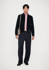 Veste texturée bleu marine, col roulé rose clair, pantalon sombre à rayures et chaussures noires élégantes. Petit sac bandoulière marron clair.