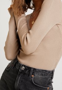 Beige geribbelde langemouwen top met een V-hals, gecombineerd met donkergrijze hoge taille jeans. Bevat een zilveren knoop en koperen accenten.