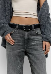 A szürke farmernadrág csillagmotívumokkal van díszítve, amelyeket strasszkövek ékesítenek, és egy fekete övvel, valamint egy testhez simuló krémszínű top alatt viselnek, amely egy szürke kötött kardigán alatt található.