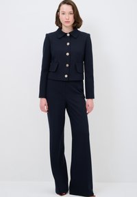 Námořnický krátký blazer s velkým límcem, zlatými knoflíky a dvěma předními kapsami v kombinaci s odpovídajícími širokými kalhotami. Hladká, dobře střižená tkanina.