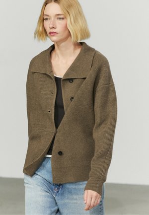 Donna bionda con un cardigan marrone lavorato a maglia con bottoni neri sopra una camicia nera e jeans azzurri, in piedi davanti a uno sfondo chiaro.