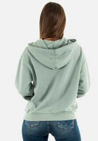 Sudadera con capucha de color verde claro y corte relajado, con puños y dobladillo acanalados. Textura suave y un notable detalle del cordón en la capucha.