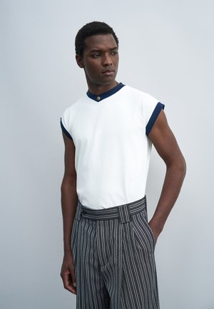 Uomo che indossa un top bianco senza maniche con rifinitura blu navy e bottone, abbinato a pantaloni grigi a righe in vita alta, in piedi su sfondo semplice.