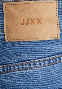 Blåt denimstof med orange syning og en rektangulær brun læderlap i midten, præget med "JJXX" i store bogstaver.