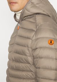 Mies, joka pukeutuu vaaleanruskeaan puhvijakkoon, jossa on huppu, leikatusta kasvoista, oranssista vetoketjun vedin ja "Save the Duck" -logopatchi hihassa.