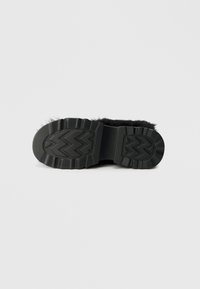 Črna čevljarska podloga s krznom in teksturirano gumijasto zunanjo podlogo, ki vsebuje zigzag vzorce za oprijem in stabilnost, s poudarkom na trajnosti in udobju.