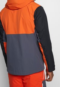 Windbreaker jack met een oranje bovenhelft, grijze middensectie en zwarte onderkant; gladde textuur, verstelbare capuchon en contrasterende mouwen.