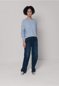 Hellblauer Strickpullover mit gerippten Bündchen, kombiniert mit leicht lockeren dunkelblauen Jeans. Das Modell trägt silberne Schuhe. Einfache Hintergrundgestaltung.