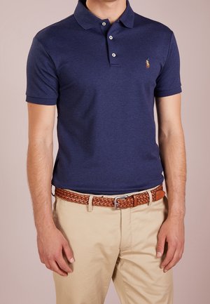 Homme portant un polo bleu marine à manches courtes et un pantalon beige avec une ceinture tressée marron sur un fond uni.