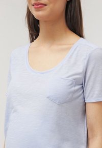 Helles blaues T-Shirt mit kurzen Ärmeln und einem Rundhalsausschnitt, das aus strukturiertem Stoff besteht und eine kleine Brusttasche hat.