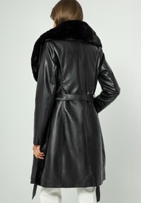 Svart läder trenchcoat med en stor krage i syntetpäls, bälte i midjan och långa ärmar. Slät textur med skräddarsydd passform och subtil lyster.