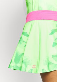 Grön klänning med utsvängd kjol, med ett rosa midjeband och subtilt ton-i-ton-mönster. Tyget är slätt med en lätt textur. Logotyp synlig.