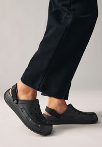 Sabots en plastique noir avec des trous de ventilation, une bride arrière et une semelle texturée, portés avec un pantalon foncé, montrant le positionnement du pied.